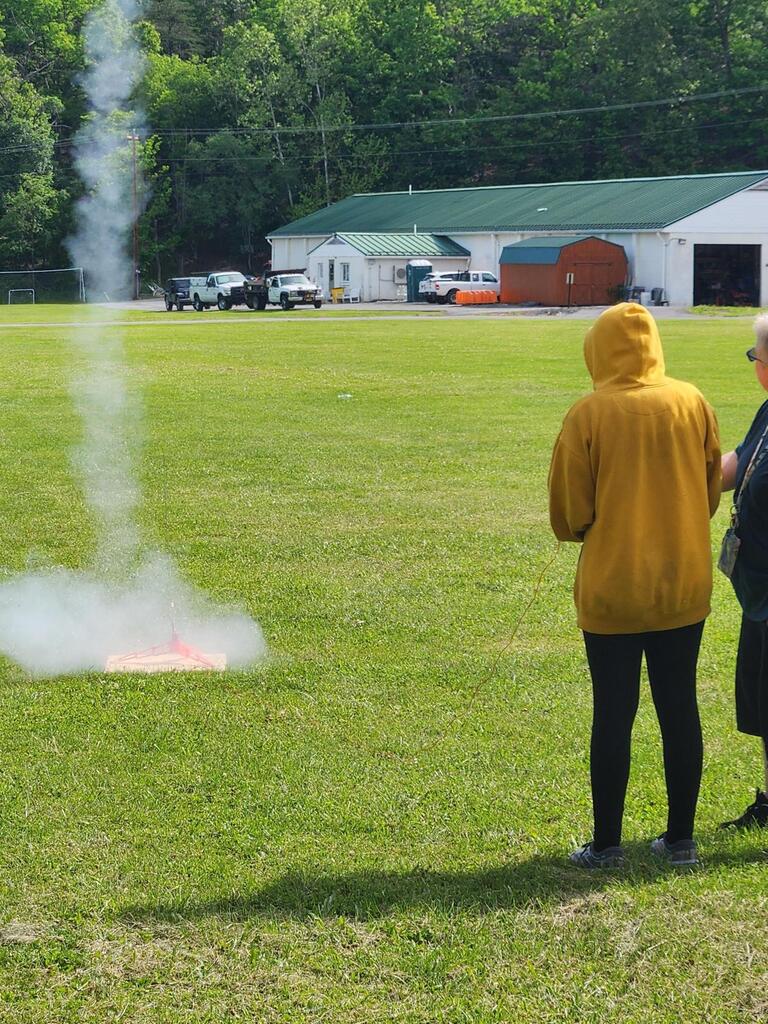 Coach Biddle and Brooklyn launch Brooklyn's rocket into the air. Smoke surrounds the launch pad after the rocket took flight.