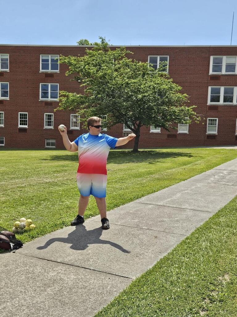 A male camp participant stands on the sidewalk between two patches of grass holds up a ball that he is getting ready to throw.