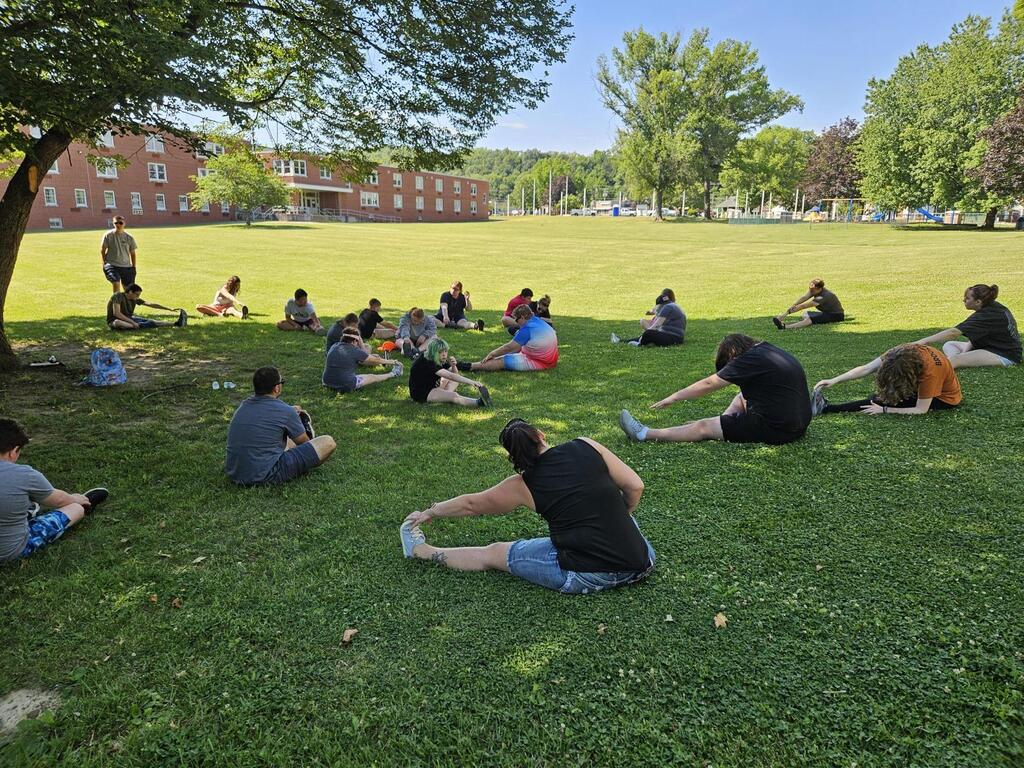 Camp participants are seated on the green grass outdoors while they stretch before competing.