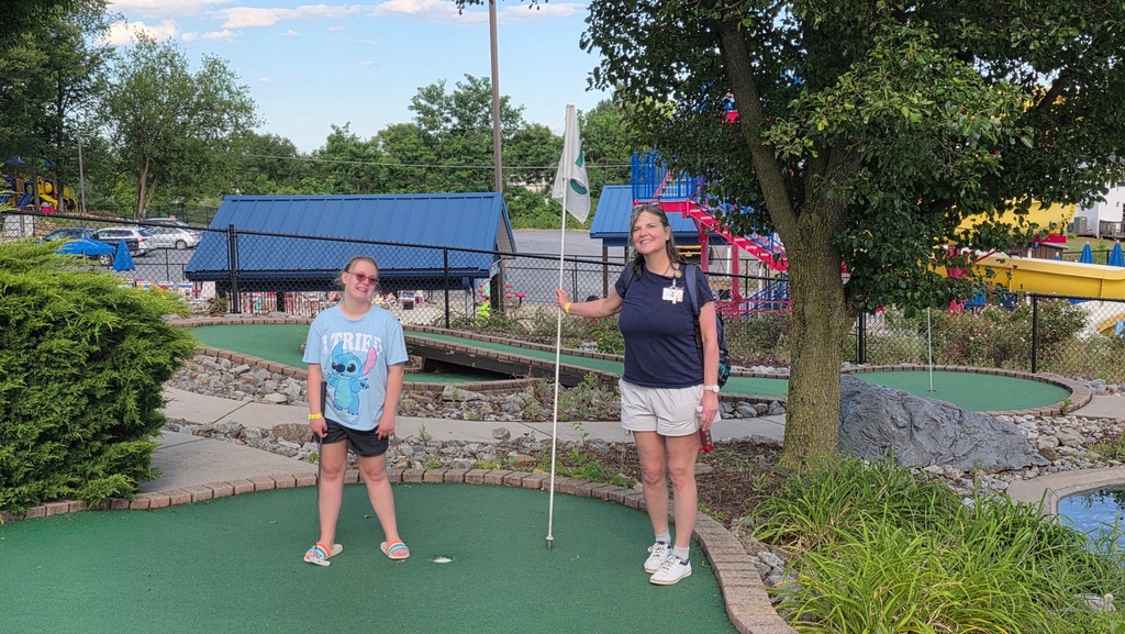 A female camp participant, Mira smiles for the camera as her and a female camp volunteer stand on the green turf of the mini-golf course.