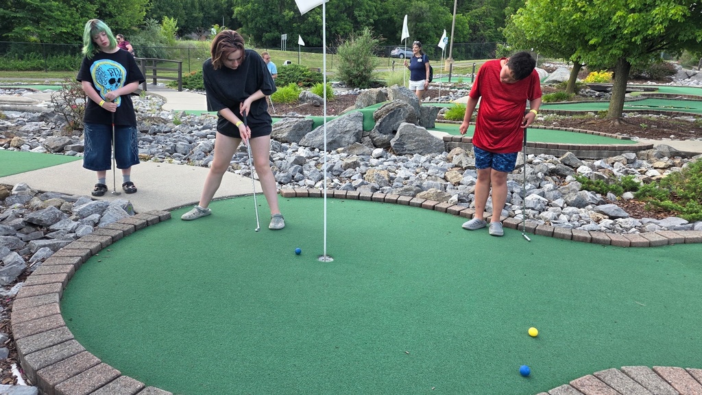 A female camp participant hits a blue golf ball while two other campers observe her.