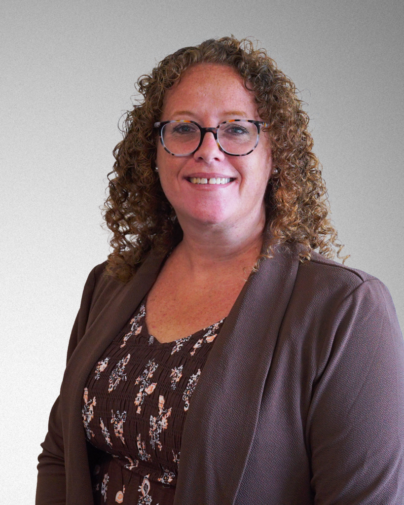 Mrs. JoAnna Gilbert. A female with brown, curly hair and wearing glasses smiles for the camera. She is wearing a black, floral blouse with a black blazer on top.