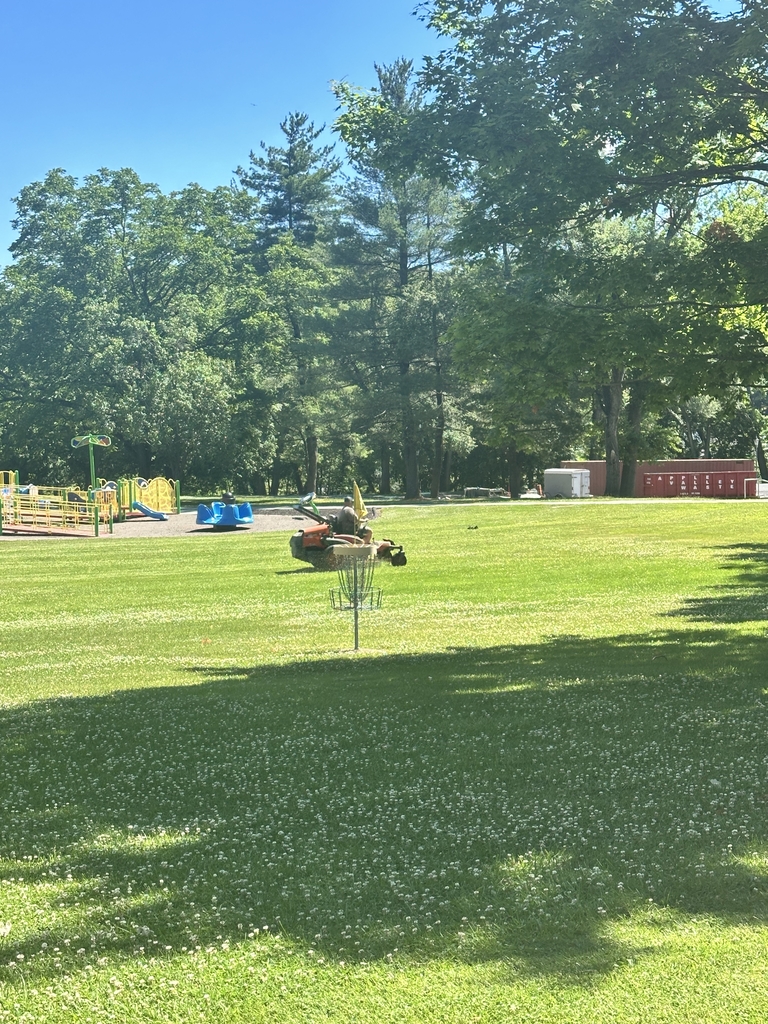 A male maintenance worker rides a lawn mower in front of a yellow and blue playground.