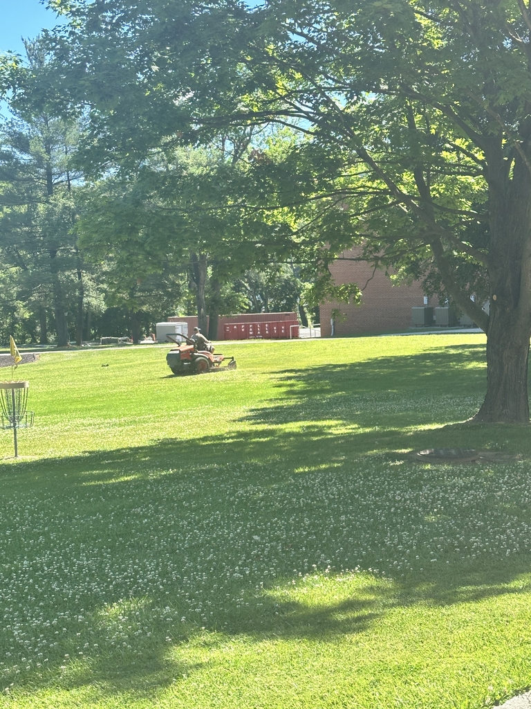 A male maintenance worker rides a lawn mower to cut the grass in front of the gymnasium.