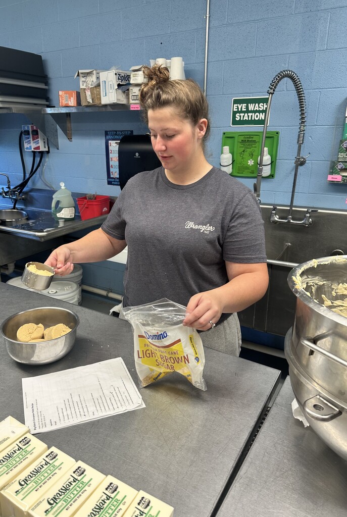 Candice, a female cafe employee looks down as she pours brown sugar from a metal measuring cup into a metal bowl.