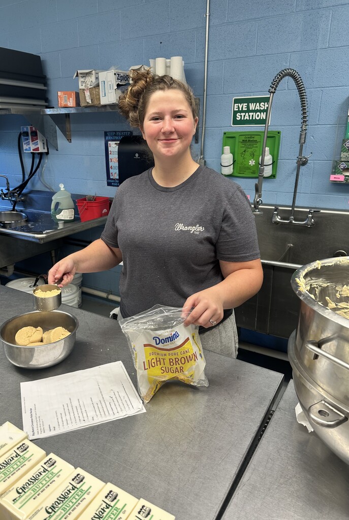 Candice, a female cafe employee smiles as she holds a measuring cup in one hand and a bag of brown sugar in the other.