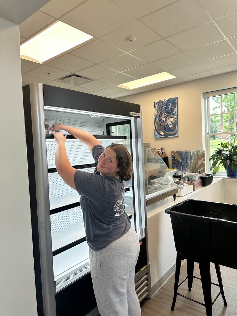 Candice, a female cafe employee looks back as she reaches the top shelf of the cooler to stock it with cafe food items and drinks.
