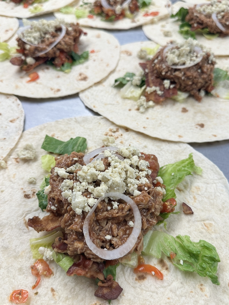 BBQ, Bacon, and Blue Chicken Wrap. Shredded BBQ chicken sits on a white tortilla and a bed of lettuce. Toppings include onion, tomatoes, and feta cheese.