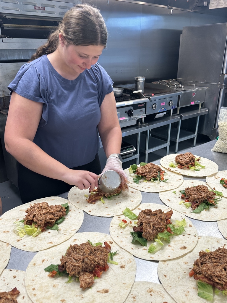 Candace, a female café worker looks down as she places BBQ chicken onto a bed of lettuce with a white tortilla underneath.