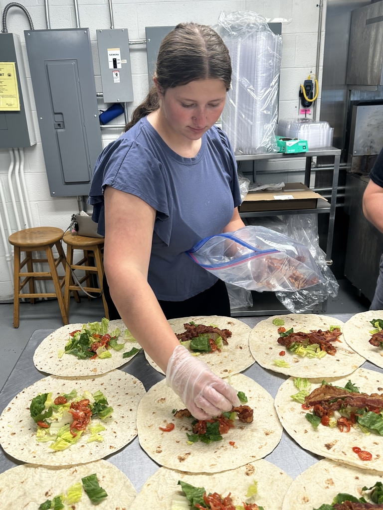 Candace, a female café worker looks down as she places pieces of bacon onto a white tortilla.