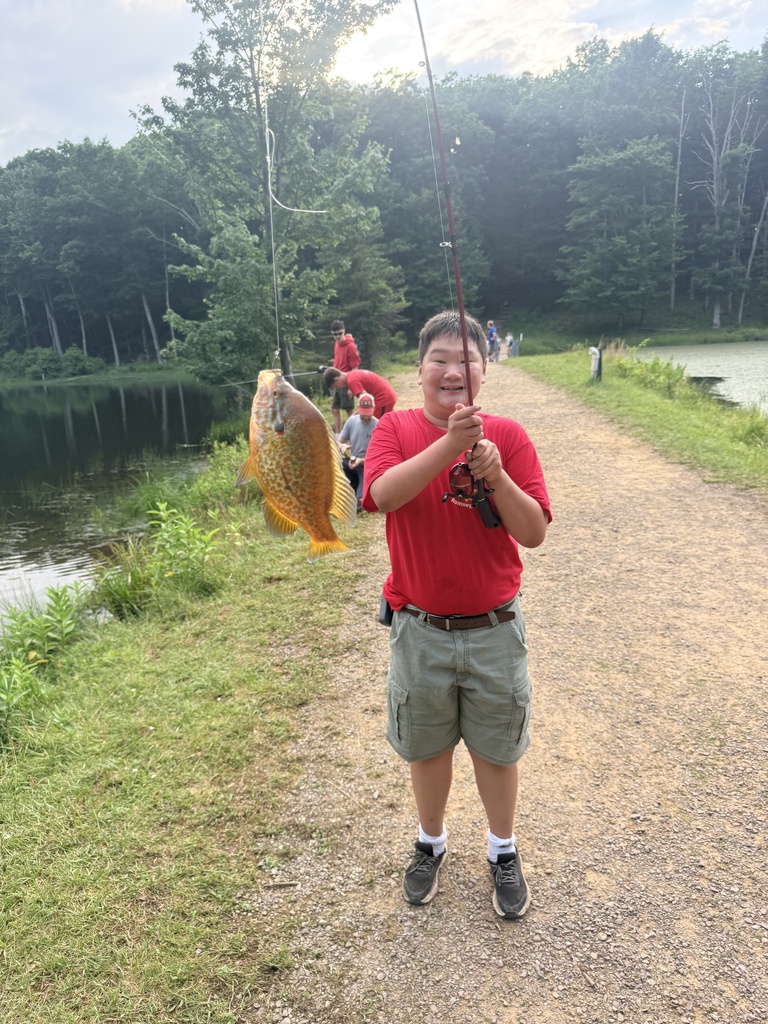 Tristian wears a red shirt and gray cargo shorts excitedly holds up a fishing pole with an orange fish hooked onto the pole.