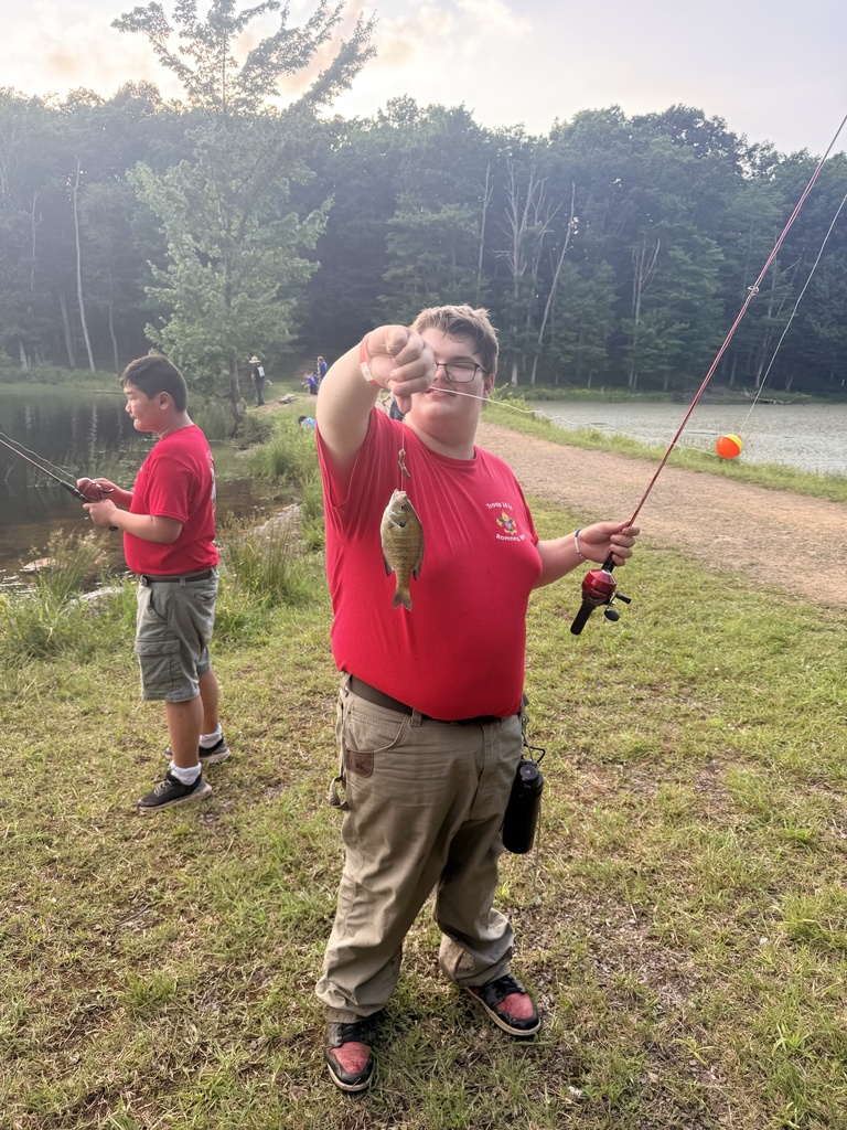 Carson, dressed in a red shirt and khaki-colored pants holds a fishing pole with a fish hanging from the hook.