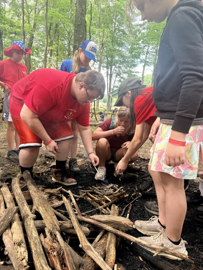 Carson dressed in a red shirt and shorts bends down as he lights a pile of wood with a match.