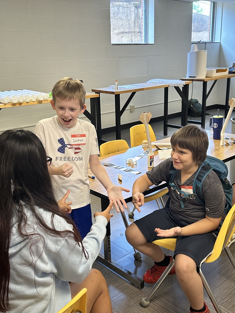 Three camp participants are gathered in a group, two are seated and one camper is standing as they play a game of rock, paper, scissors.