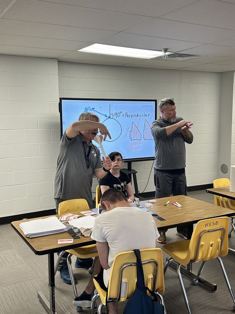 Two camp participants are seated at a rectangular table as Dr. Ensign stands at the front of the room demonstrating a hands-on learning lesson. There is a male interpreter to the left of Dr. Ensign.