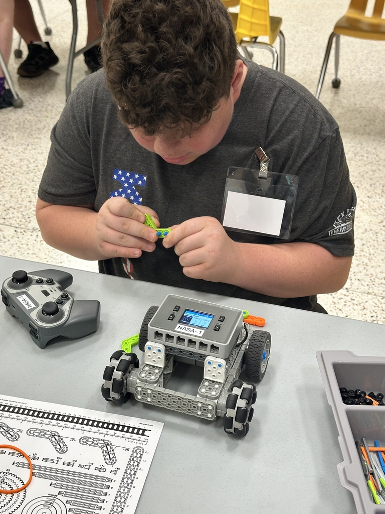 A male camp participant looks down towards a gray-colored VEX robot that is placed in front of him as he holds a green and blue-colored part that attaches to the robot.
