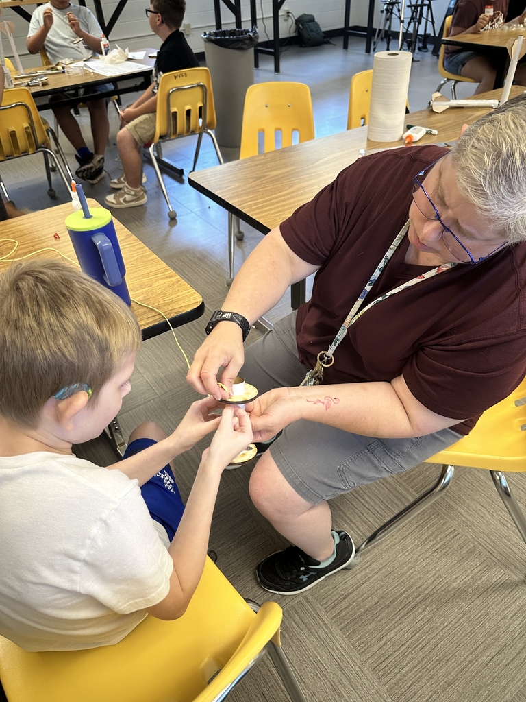 Ms. Biddle, a WVSDB educator, assists a male camp participant with putting a piece of rope into a wooden, circular vessel.