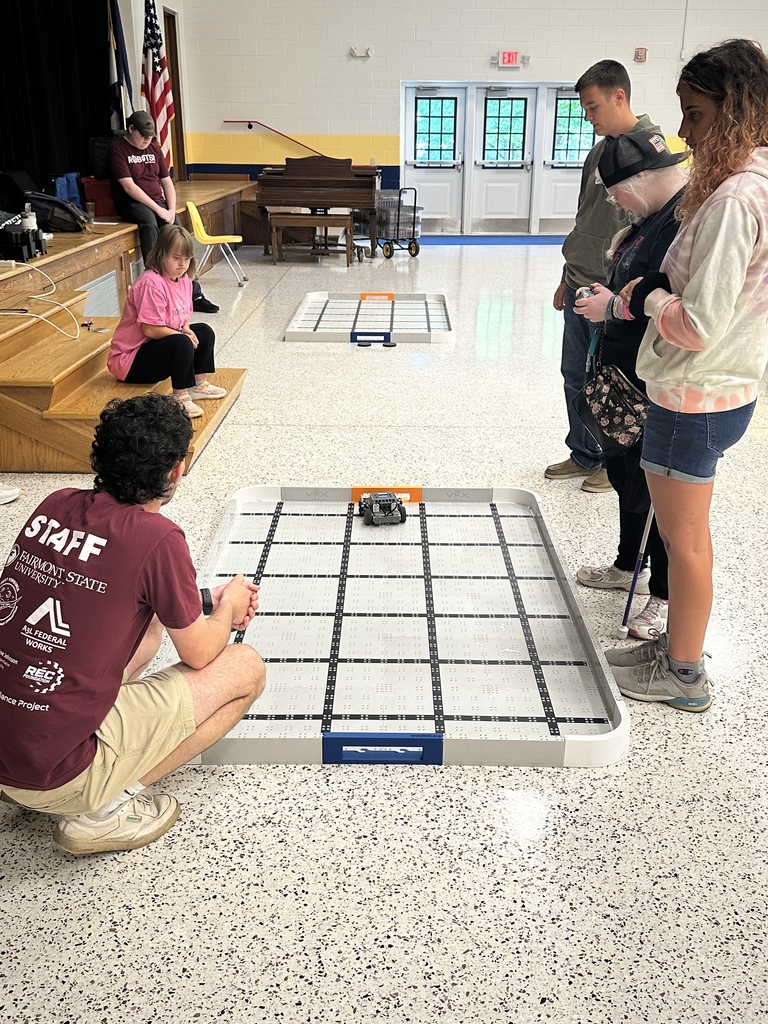 Camp participants are gathered around a VEX robot arena as they observe a female camp participant control the robot using a remote controller.