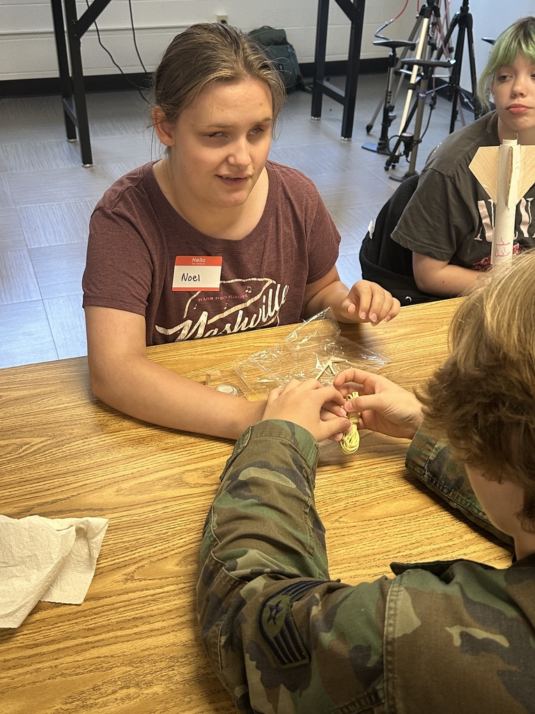 A female camp participant smiles as she has the assistance of a male NASA representative with touching the piece of rope they are going to use for their rocket vessel.