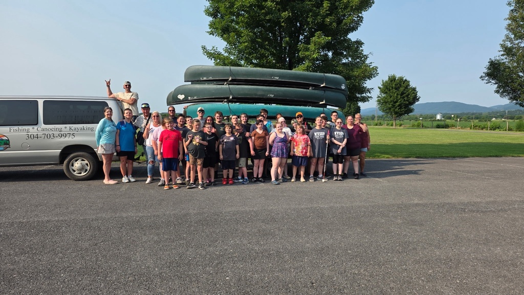 Group photo of summer camp participants and Breezewood Adventures employees standing in front of a gray van that is hauling kayaks and canoes.
