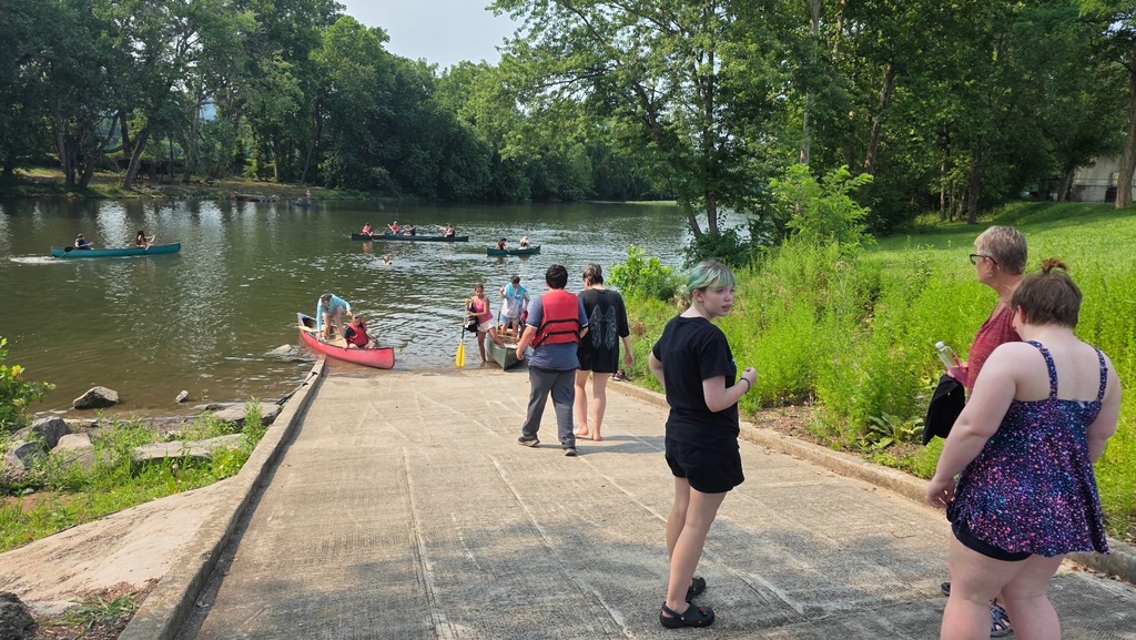 Camp participants make their way down to the riverbank to load up into their canoe/kayak. There are multiple canoes/kayaks already floating in the water in the background.