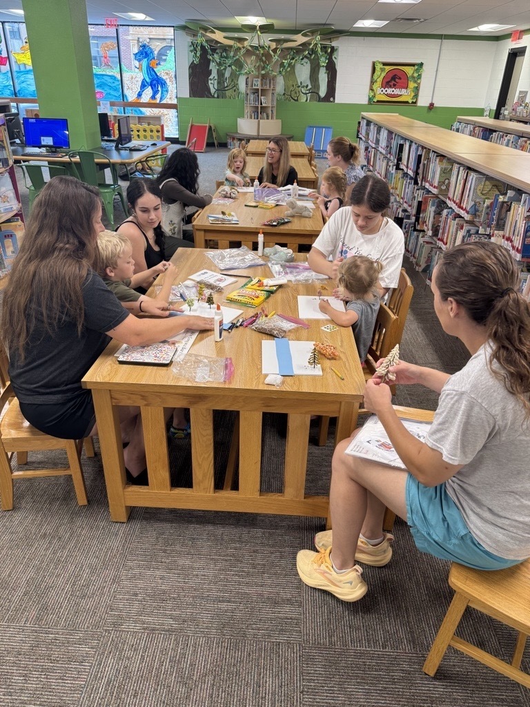 Children and their families are seated at wooden tables working on numerous crafts. Craft supplies are spread across the table.