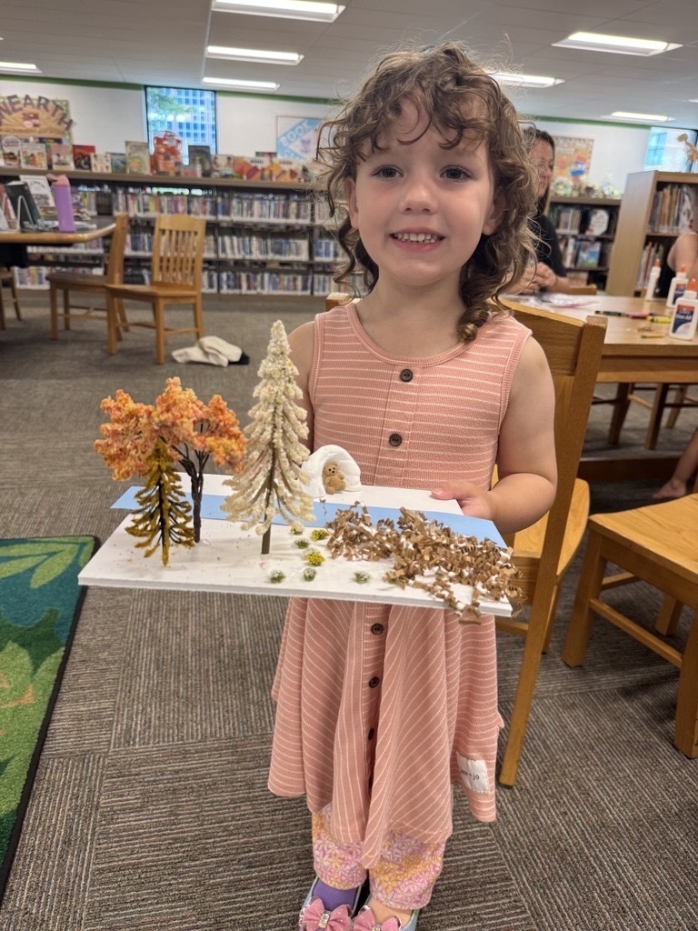 A little girl wearing a pink and white striped dress smiles excitedly while she holds up a craft, she created that consists of a seasonal changes.