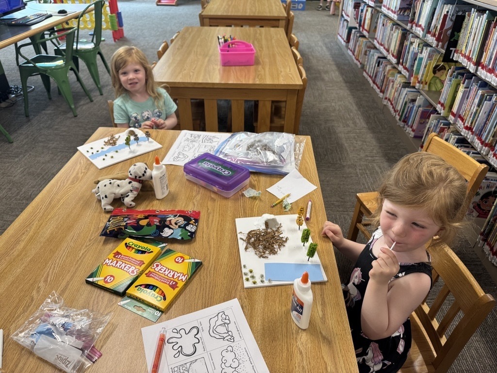 Two children are seated at a wooden table with craft supplies spread across the table.
