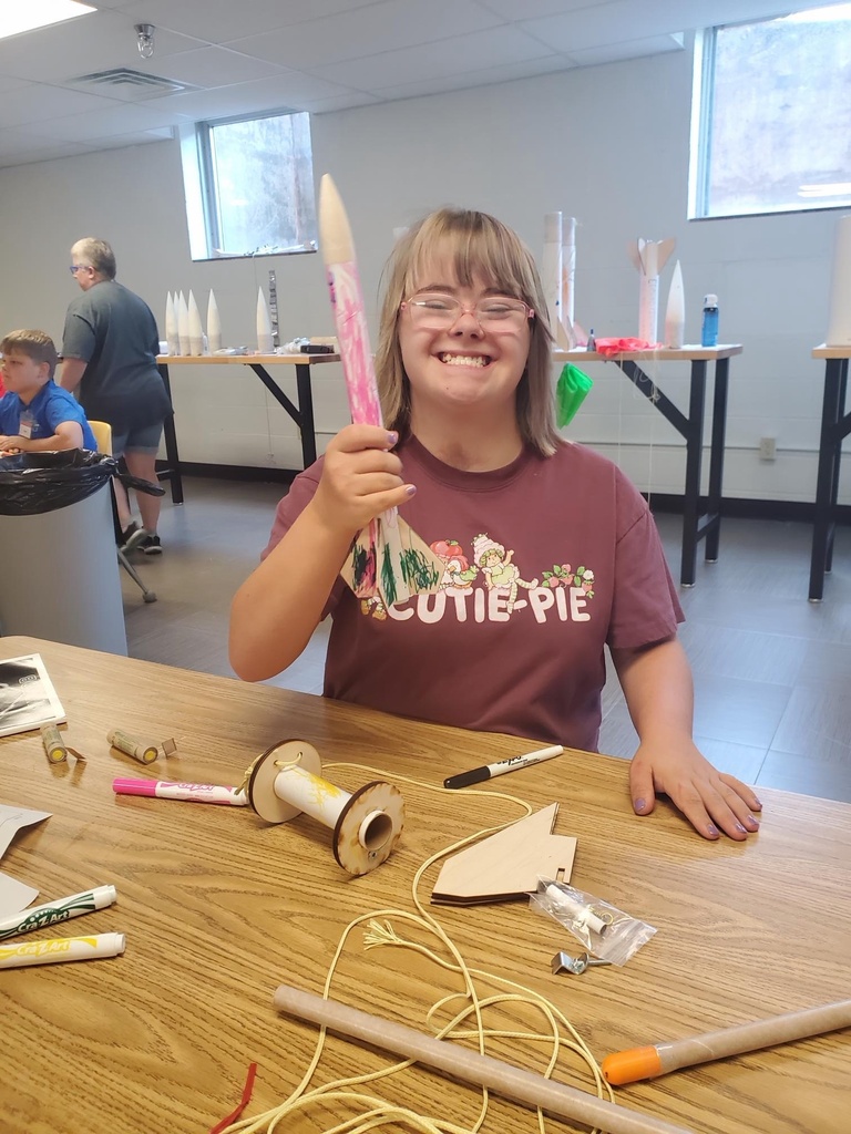 A female camp participant with blonde hair and wearing glasses excitedly smiles and holds up the rocket she built.
