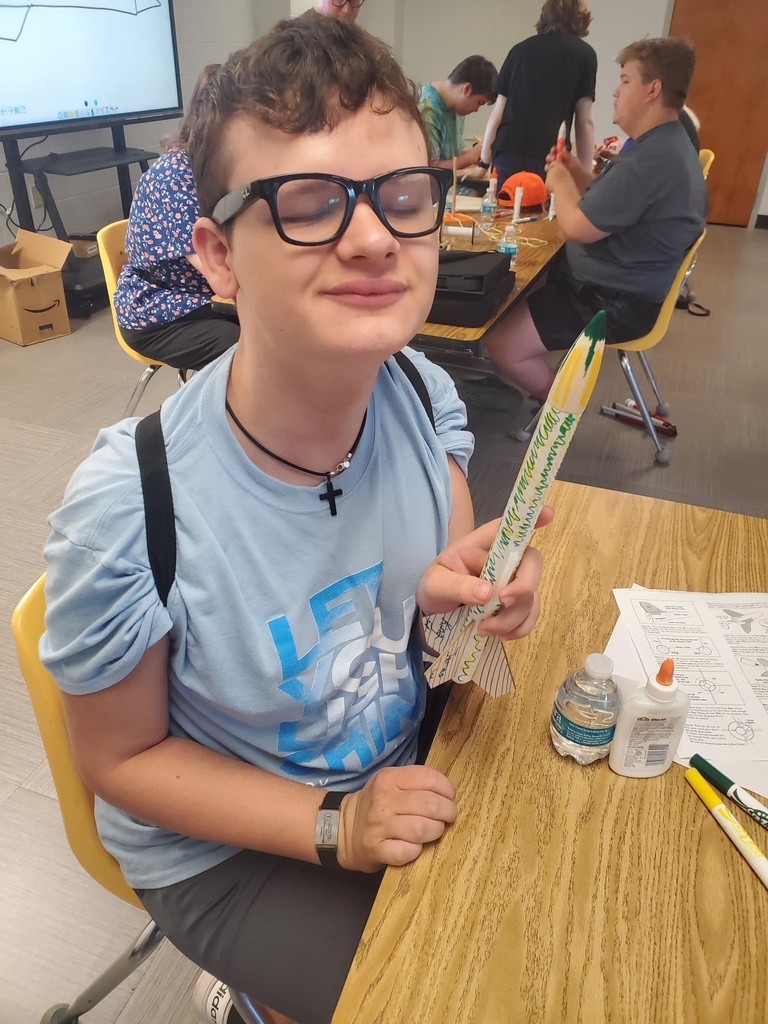 A male camp participant wearing a blue shirt and wearing glasses holds up the rocket he built.