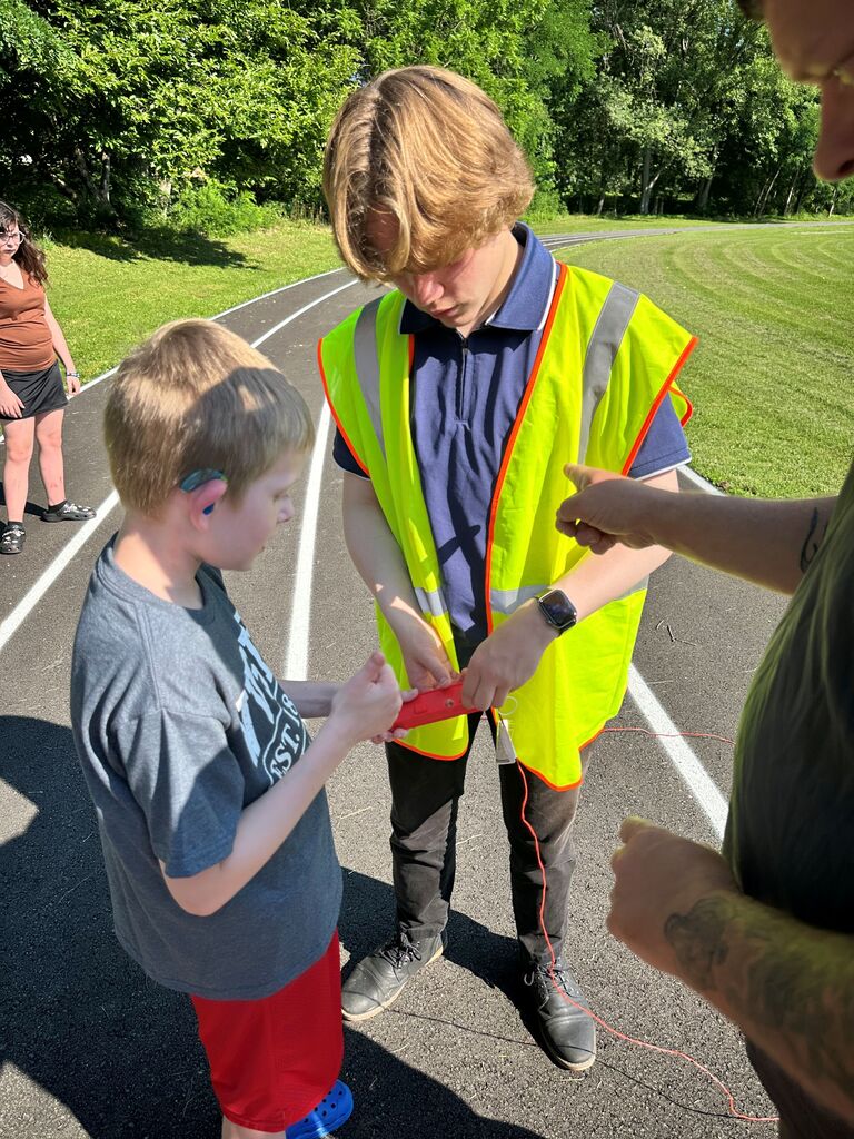 A male camp volunteer wearing a bright, yellow vest assists a male camp participant with pressing the button to launch his rocket.