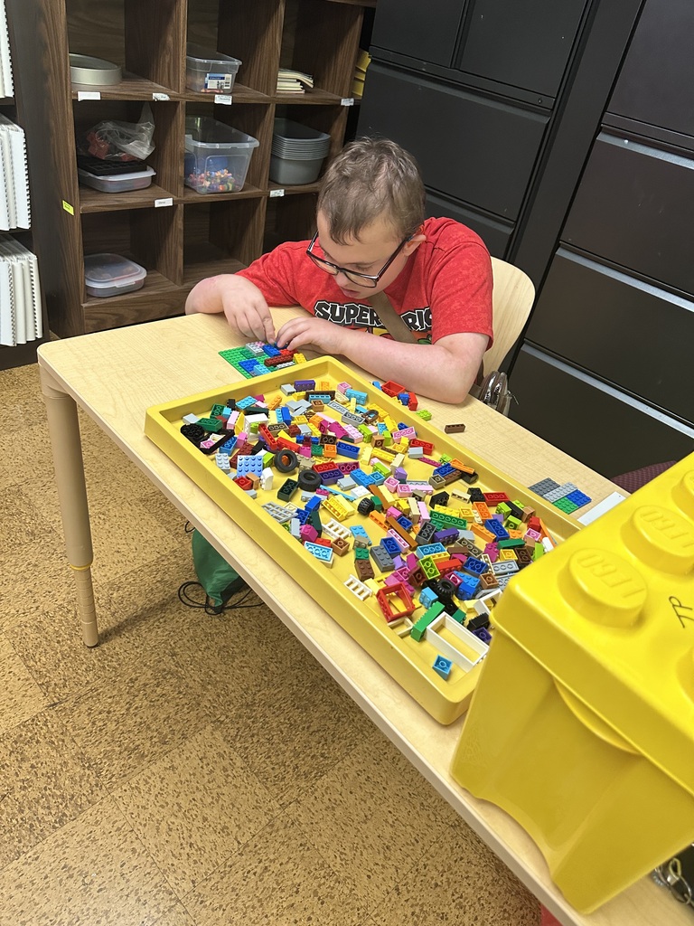 A male camp participant looks down as he puts together Lego pieces. There is a large yellow box full of Legos sitting beside him.