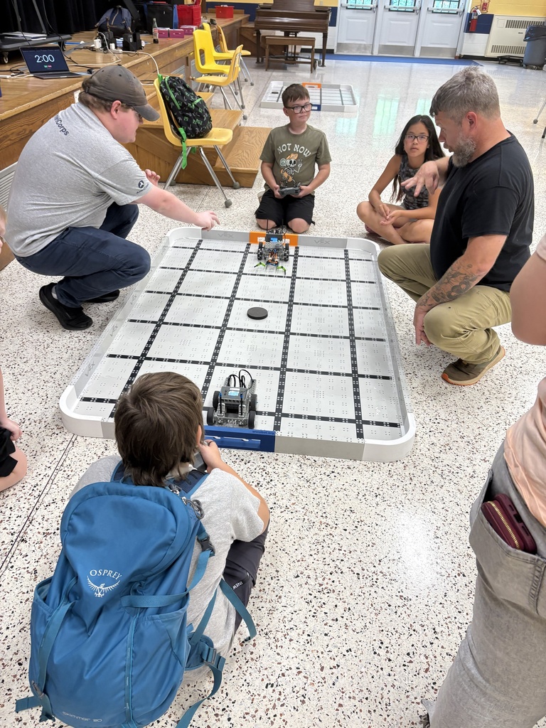 Camp participants and volunteers are gathered around a gray VEX robot arena with two robots inside of the arena.