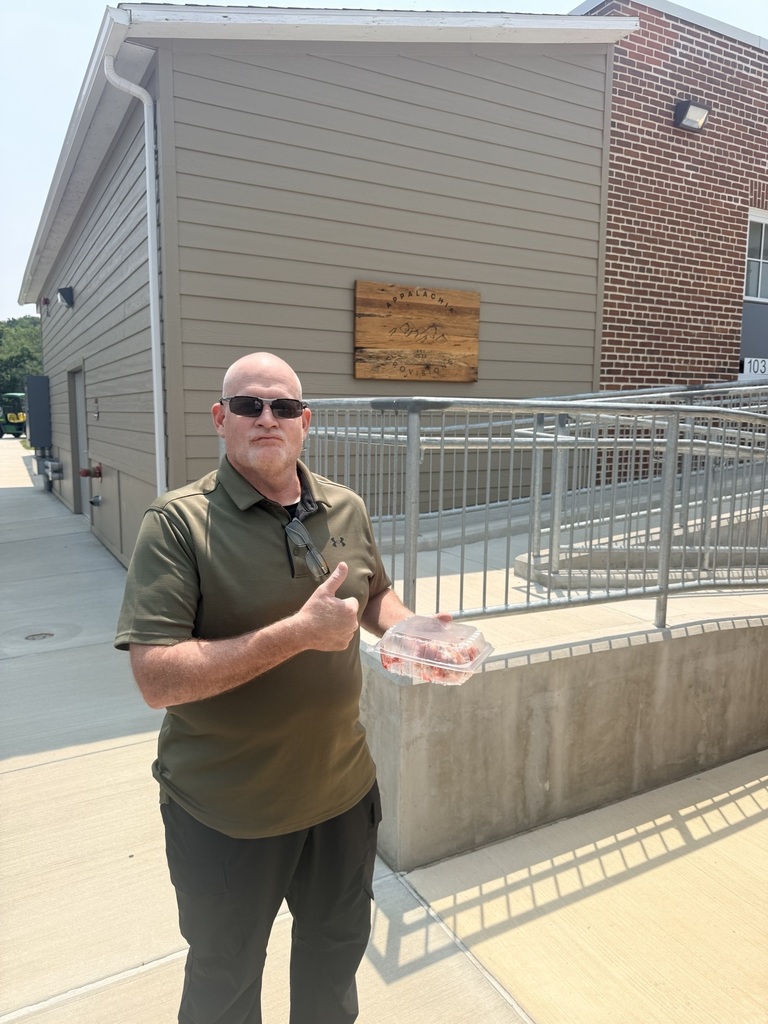 A visitor gives a thumbs-up while holding a dessert outside Appalachia Provisions Café in front of the café's wooden sign and accessible entrance.