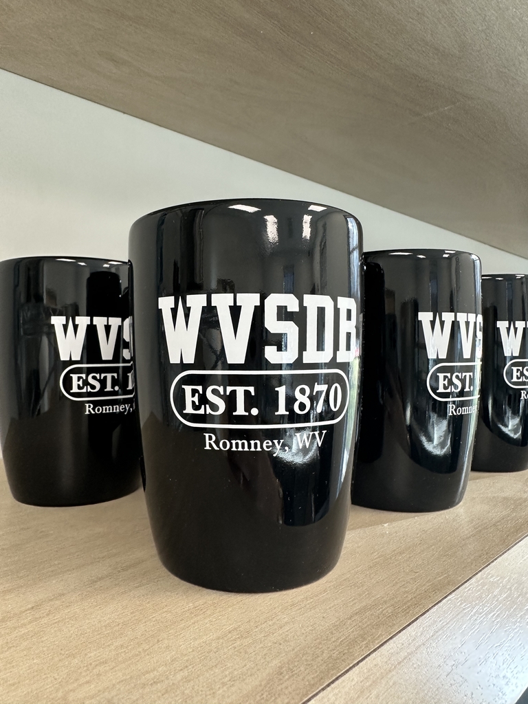 Black WVSDB ceramic coffee mugs displayed on a wooden shelf, featuring a white "WVSDB Est. 1870 Romney, WV" design.