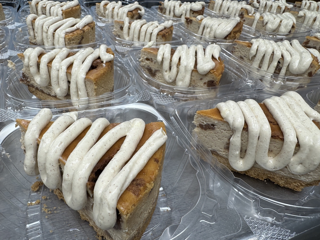 Individual slices of cinnamon roll cheesecake topped with a thick cinnamon cream drizzle in clear dessert containers.