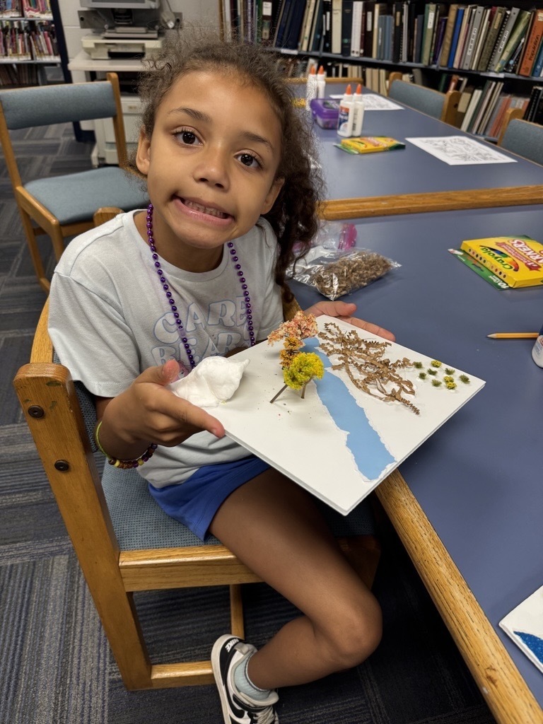 A smiling student holds up a completed nature-inspired art project featuring dried flowers, leaves, and a blue paper design while sitting at a table in a library classroom with craft supplies nearby.