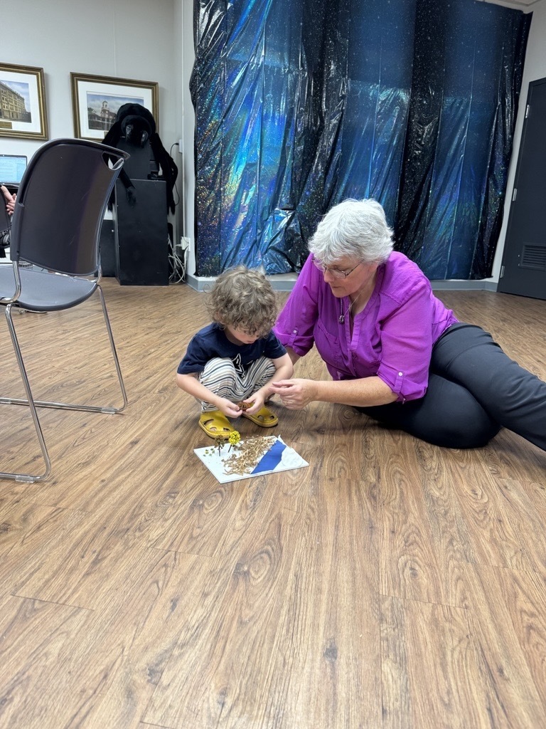 An adult sits on the floor beside a young child, helping them arrange dried flowers, leaves, and other natural materials on a small art project. Both are focused on the activity.