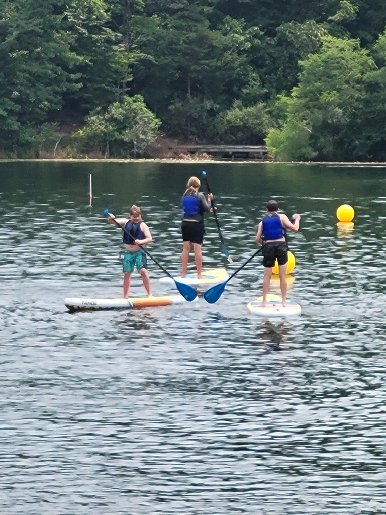 Three WVSDB Boy Scouts paddle board across a calm lake while wearing life jackets during the National Scout Jamboree.
