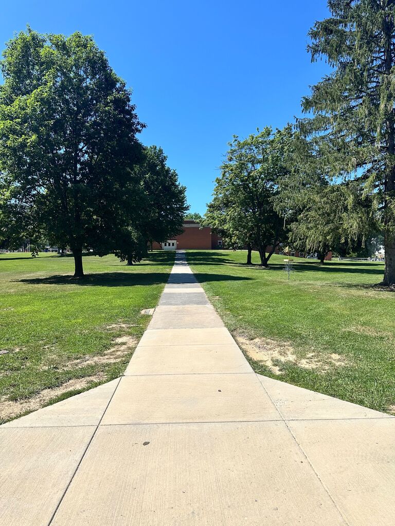 Concrete sidewalk leading through a green campus lawn toward the Sevigny building, bordered by trees on a bright summer day.