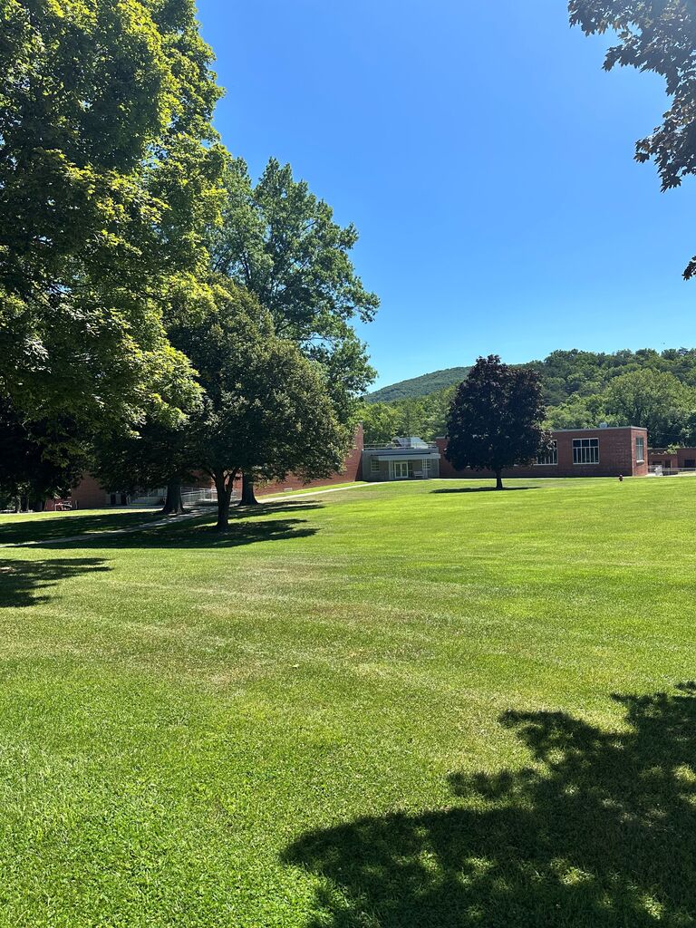Wide view of the WVSDB campus on a sunny summer day, featuring a large green lawn, mature shade trees, the PE building, and forested mountains beneath a clear blue sky.