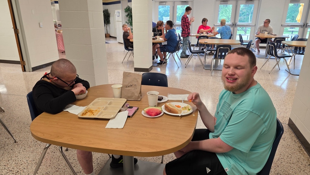 Two students enjoy lunch together at a table in the campus dining hall while other students and staff eat and socialize in the background.