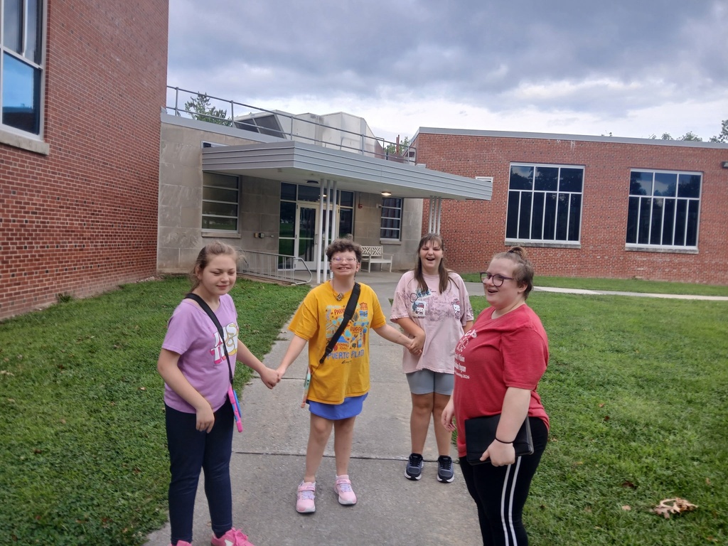 Four students stand outside on a campus walkway holding hands and smiling near the residence hall on a cloudy day, enjoying time together between activities.