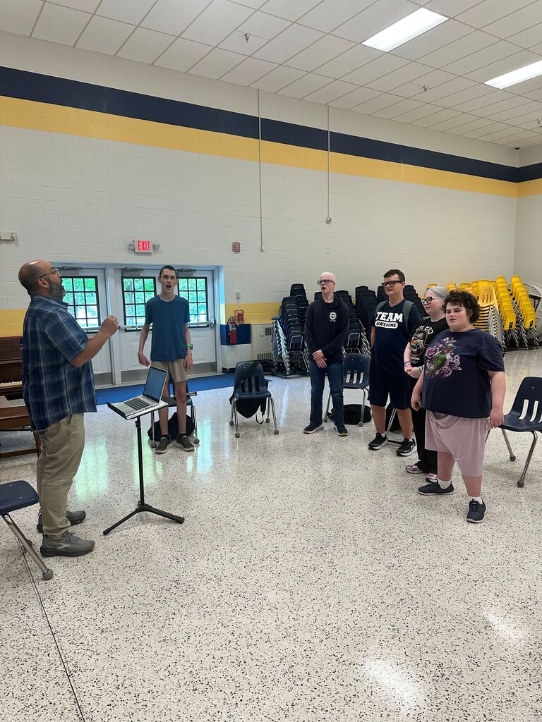 Music teacher leads a small group of students in choir rehearsal inside a large multipurpose room. Students stand in a semicircle singing while the teacher conducts beside a piano and laptop.