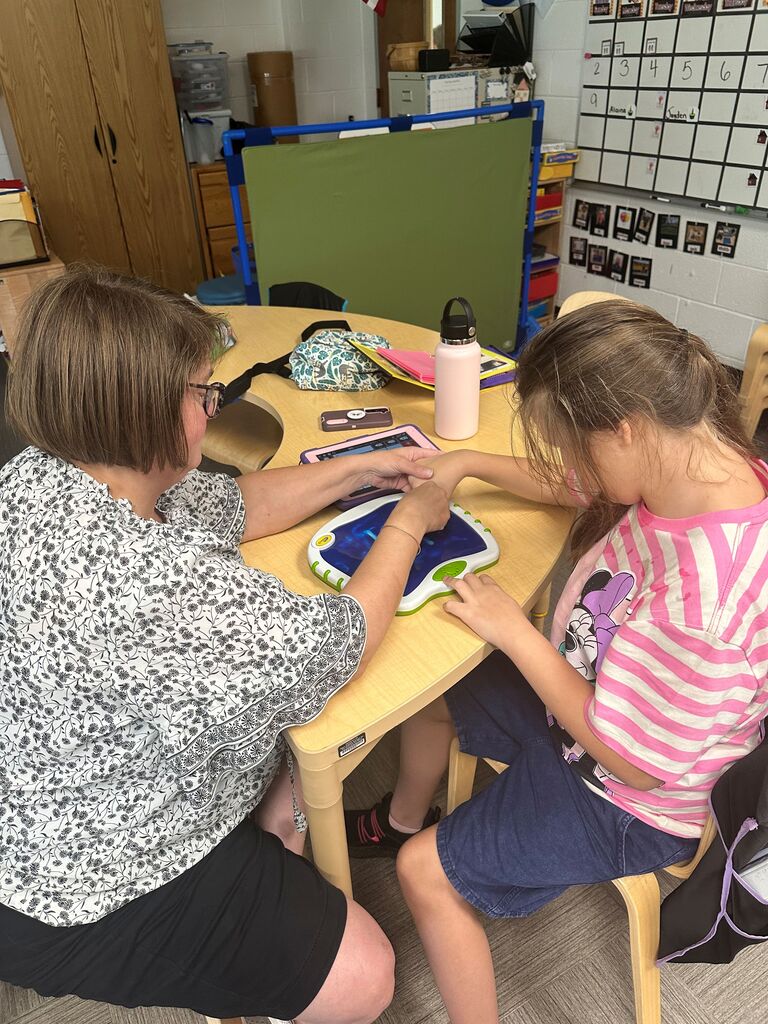 Teacher works one-on-one with a young student using a digital display at a classroom table, guiding the student's hands during a literacy activity.