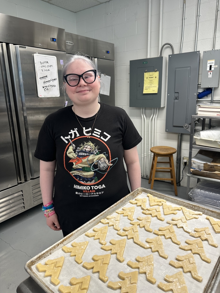 Student stands proudly in the culinary classroom beside a baking tray filled with uncooked WVU-themed cookies, ready for the oven.