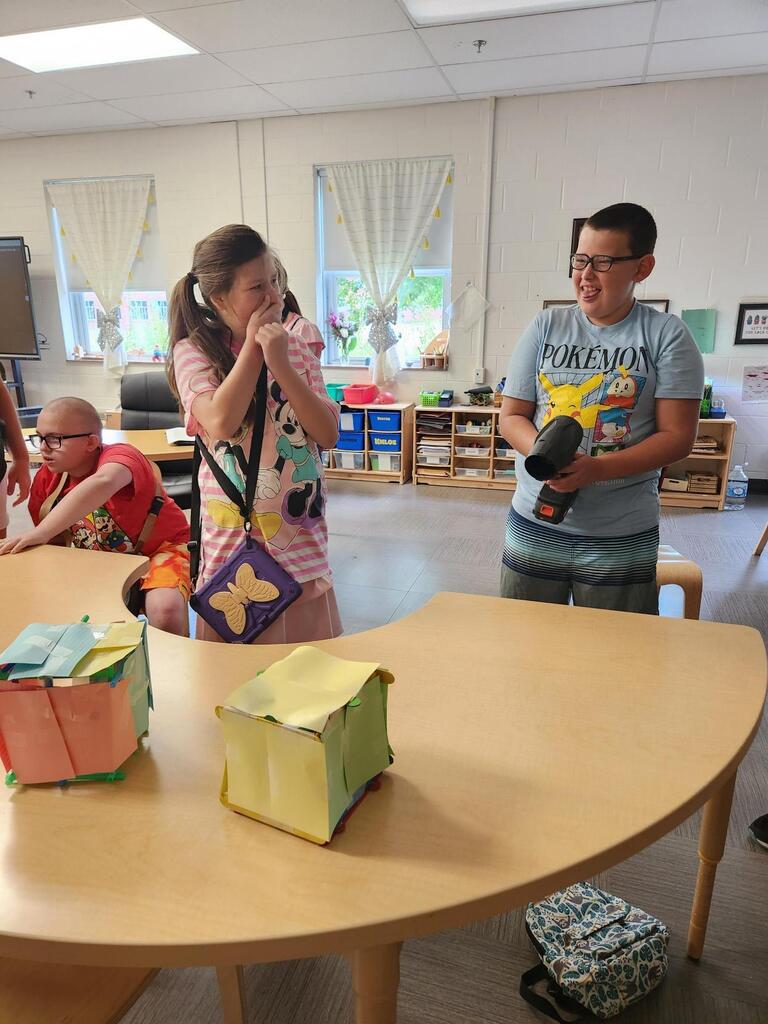 Two students smile and react as one uses a handheld blower to test the effects of strong winds on colorful model structures during a hurricane science activity.