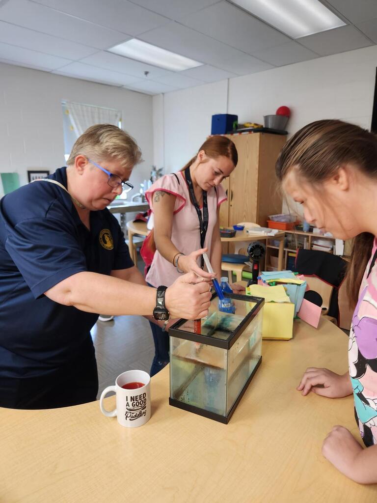 A teacher demonstrates a water temperature experiment using a small aquarium while two students watch closely. Colored water in the tank helps illustrate how warm and cold water move and interact.