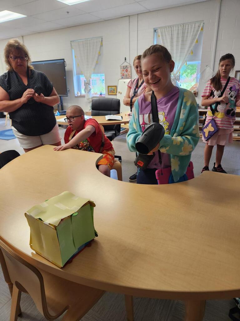 A smiling student uses a handheld blower to test how a student-built model structure responds to strong winds while classmates and staff watch.
