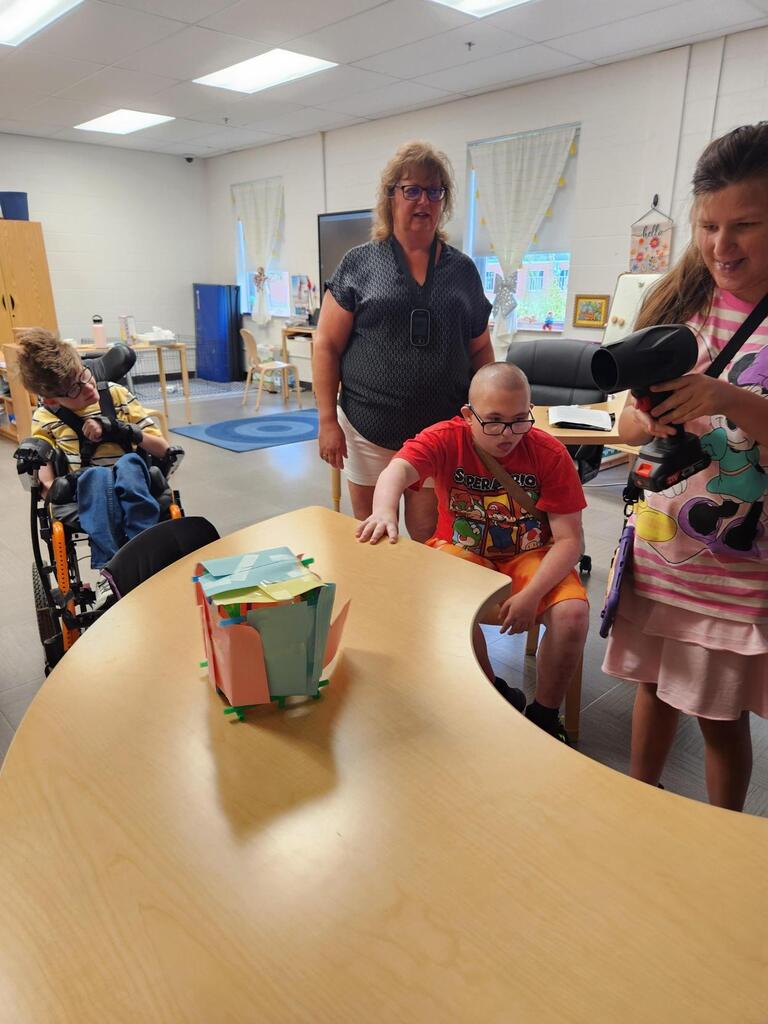 Students and staff gather around a table as a student uses a handheld blower to test a colorful model structure against strong winds during a hands-on hurricane science lesson.